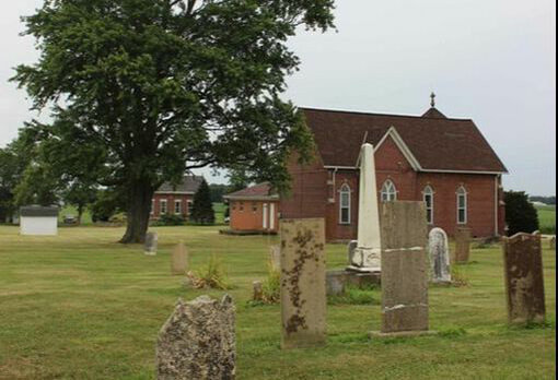 grave markers in front of a red brick church building with a tree on the side